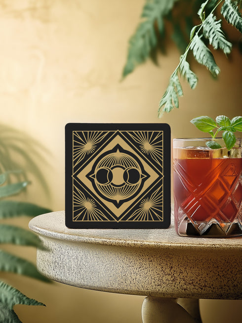 A black and gold ceramic coaster featuring a triple moon design framed by art deco geometric details and celestial sunburst accents.